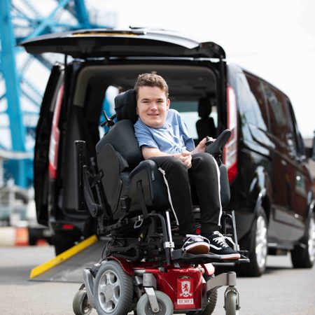 A boy in a wheelchair is next to a car. 