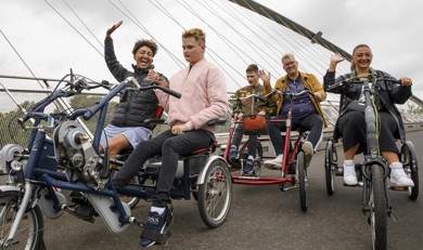 Three inclusive cycles are pictured on a bridge. The first seats two young men one has brown hair, black coat, and blue shorts, the other has blond hair, pink jacket, black jeans, and black trainers. Another cycle has a young man with brown hair in a green jacket with badges, black trousers and blue trainers and a man with white hair, yellow jacket, blue jumper, black trousers, and white trainers. The final cycle is a one-person cycle, a young woman is sat with brown long hair, black jacket, black trousers.
