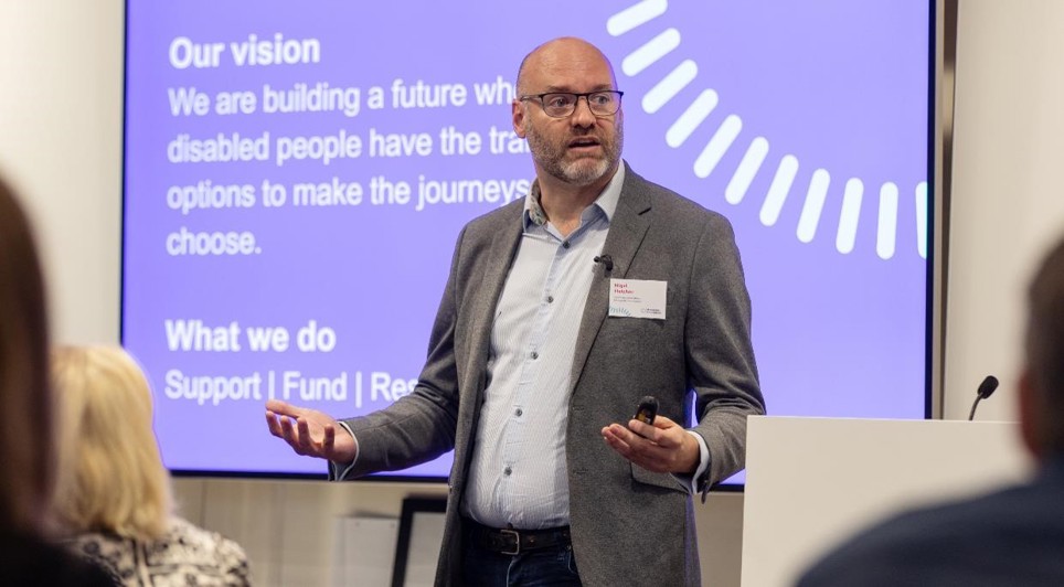 Nigel is presenting to an audience at an event. He is wearing black glasses, grey jacket and a blue shirt. 