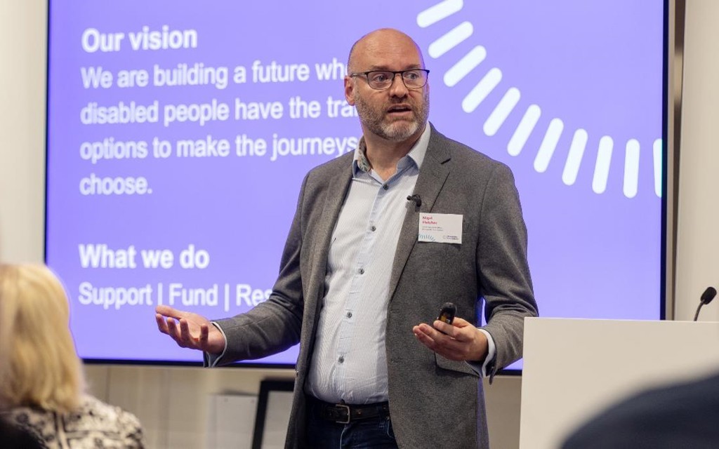 Nigel is presenting to an audience at an event. He is wearing black glasses, grey jacket and a blue shirt. 