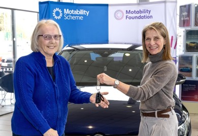 Wera Hobhouse MP presents keys to Rose at car handover in Bath. Rose's car is behind them. Rose is on the left-hand side in the photo she has white hair in a bob with glasses. She is wearing a midnight blue zip up jumper. Wera is on the left-hand side of the photo with shoulder length blonde brown hair and a grey turtleneck jumper. They are smiling looking at the camera. 