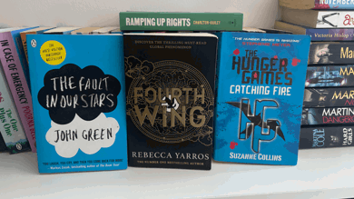 The photo is of a white bookshelf with books pilled high. Three books are displayed up right "The fault in our stars", "Fourth wing" and The hunger games catching fire".