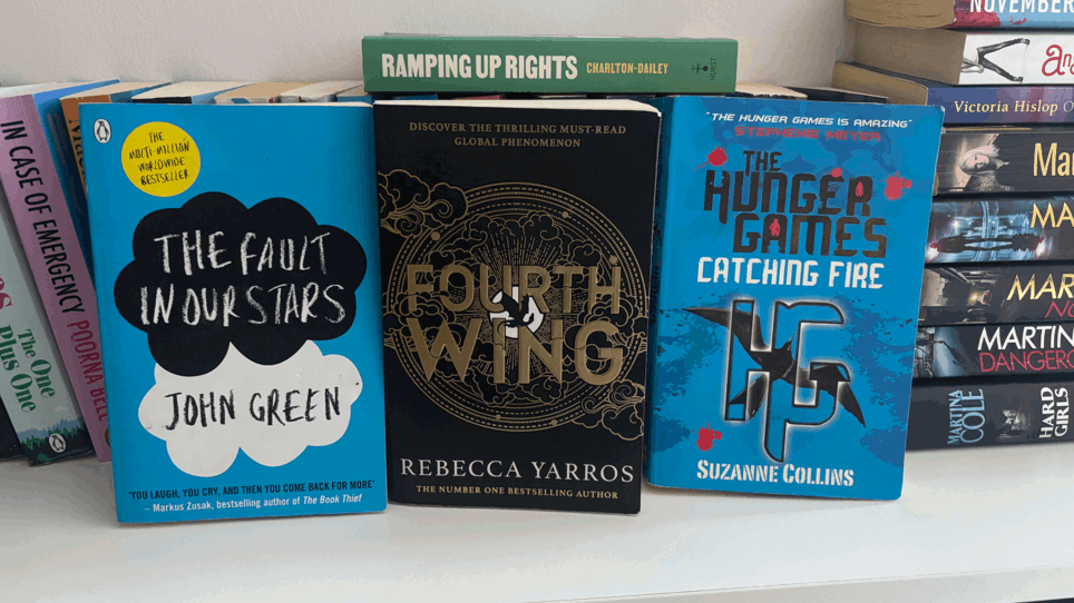 The photo is of a white bookshelf with books pilled high. Three books are displayed up right "The fault in our stars", "Fourth wing" and The hunger games catching fire".