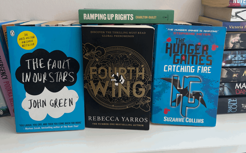 The photo is of a white bookshelf with books pilled high. Three books are displayed up right "The fault in our stars", "Fourth wing" and The hunger games catching fire".