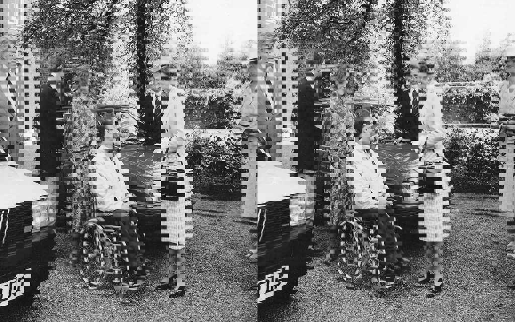 Her Majesty The Queen standing in front of a car and talking to a gentleman who is sitting in a wheelchair. They are both smiling.