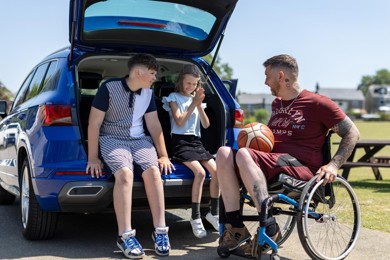 Aaron is sitting in his wheelchair next to his car. The car boot is open and his children are sitting in it. They are all talking to one another.