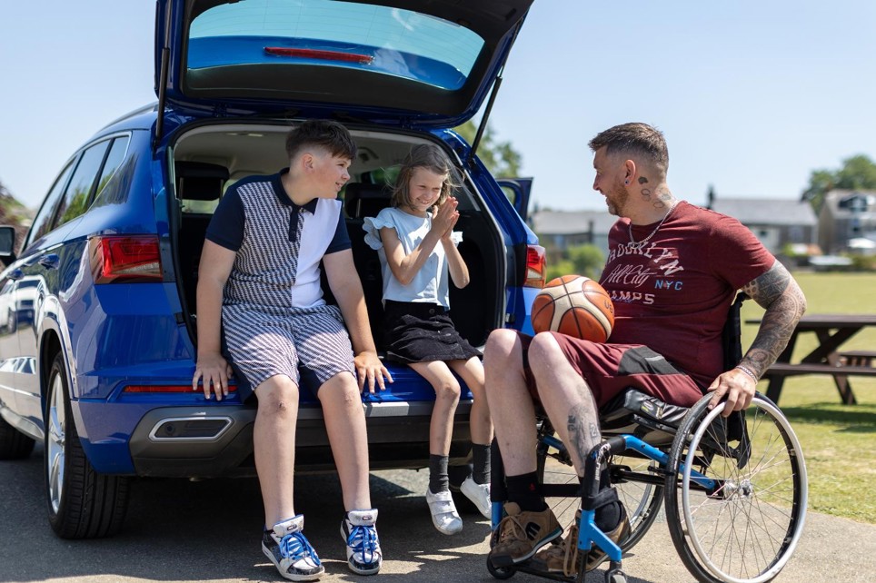 Aaron is sitting in his wheelchair next to his car. The car boot is open and his children are sitting in it. They are all talking to one another.