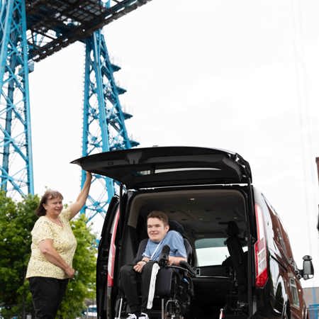 A boy in a wheelchair is driving down the ramp of a car. While a lady holds open the car door. 