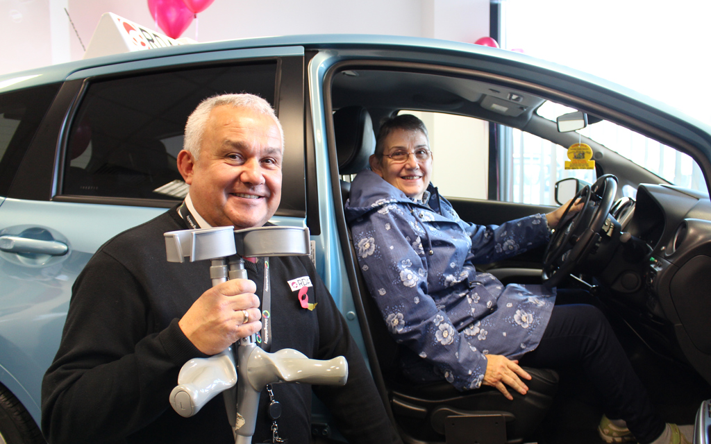 A man holding his silver crutches is standing smiling in front of a light blue vehicle. The door of the vehicle is open and a lady is sitting in the front seat smiling.