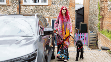 Charlie has pink hair and is wearing multi coloured clothing. They are holding their dog by the lead. Their dog is black poodle cross and is wearing a red neckerchief. Together they are on their drive next to Charlie's silver car.