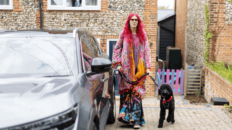 Charlie has pink hair and is wearing multi coloured clothing. They are holding their dog by the lead. Their dog is black poodle cross and is wearing a red neckerchief. Together they are on their drive next to Charlie's silver car.