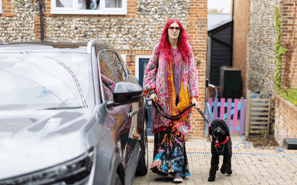Charlie has pink hair and is wearing multi coloured clothing. They are holding their dog by the lead. Their dog is black poodle cross and is wearing a red neckerchief. Together they are on their drive next to Charlie's silver car.