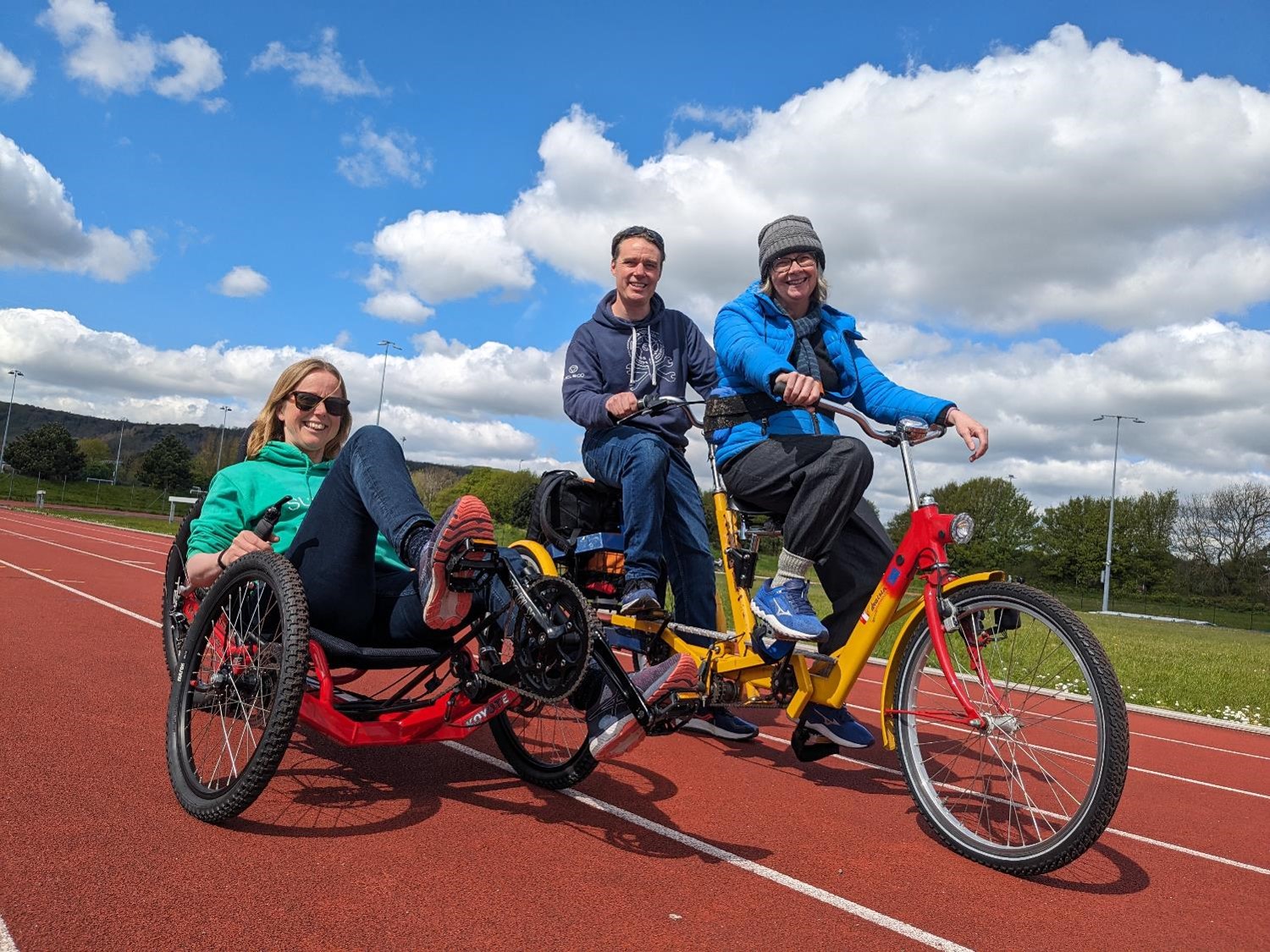 A lady is smiling while sitting in an adapted wheel on a running track with a lady and man smiling next to her on a tandem cycle. 