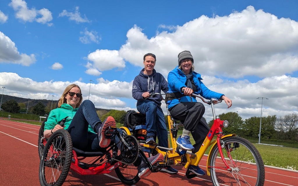 A lady is smiling while sitting in an adapted wheel on a running track with a lady and man smiling next to her on a tandem cycle.