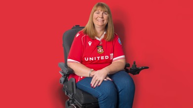 Kerry Evans is a wheelchair user. She is facing the camera smiling, she has shoulder length brown hair with a fringe. She is wearing Wrexham AFC top and blue jeans. 