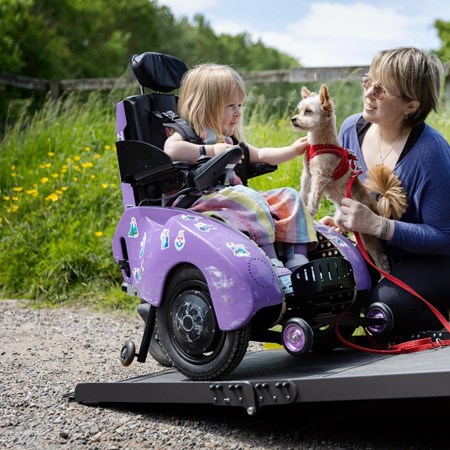 Poppy is sitting in her powered wheelchair on the ramp of her vehicle. Her mum Kim is crouched next to her, holding their family dog. Poppy is petting the dog and smiling.