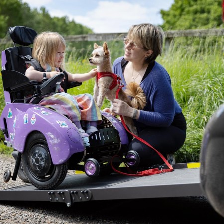 Poppy is sitting in her powered wheelchair on the ramp of her vehicle. Her mum Kim is crouched next to her, holding their family dog. Poppy is petting the dog and smiling.