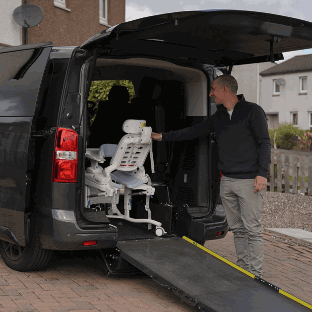 Gino's dad is wheeling a shower chair into the back of Gino's WAV, the back of the WAV is open and the ramp down. His dad is stood to the side of the ramp. 
