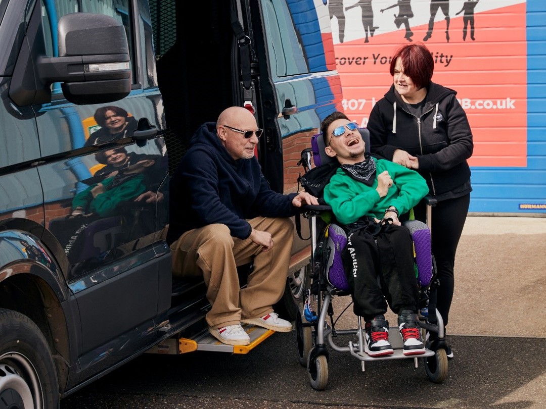 The photo is taken outside. A man is sitting on the step of the open door of a black vehicle to the left-hand side. In the middle of the photo is a young man smiling. He is sitting in his wheelchair. To the right-hand side of the image is a woman, she is looking at the man. Those photographed are Warren, Tyler and Teresa.