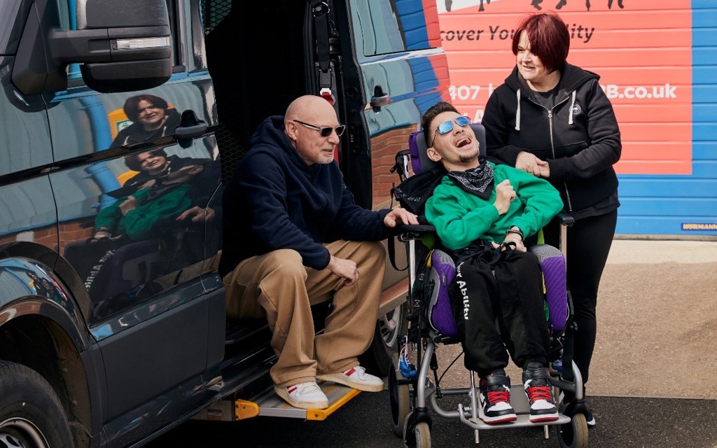 The photo is taken outside. A man is sitting on the step of the open door of a black vehicle to the left-hand side. In the middle of the photo is a young man smiling. He is sitting in his wheelchair. To the right-hand side of the image is a woman, she is looking at the man. Those photographed are Warren, Tyler and Teresa.