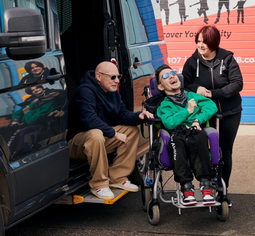 The photo is taken outside. A man is sitting on the step of the open door of a black vehicle to the left-hand side. In the middle of the photo is a young man smiling. He is sitting in his wheelchair. To the right-hand side of the image is a woman, she is looking at the man. Those photographed are Warren, Tyler and Teresa.