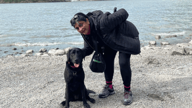 Debs is bent down in the photograph with her hand behind Betty the black labradors head. They are both looking at the camera. Behind them is a stone beach and mountain range.