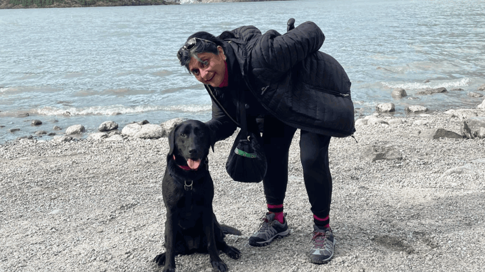 Debs is bent down in the photograph with her hand behind Betty the black labradors head. They are both looking at the camera. Behind them is a stone beach and mountain range.
