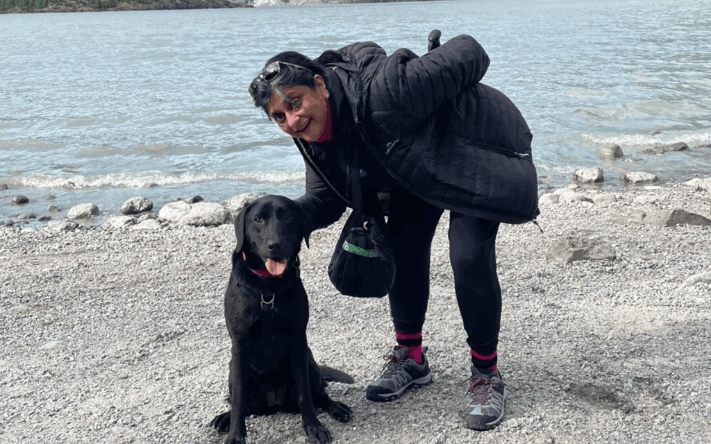 Debs is bent down in the photograph with her hand behind Betty the black labradors head. They are both looking at the camera. Behind them is a stone beach and mountain range.