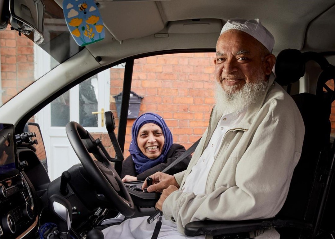 Moosa is sitting in his vehicle with hand controls smiling. His wife is behind the car, standing next to Moosa, smiling. 
