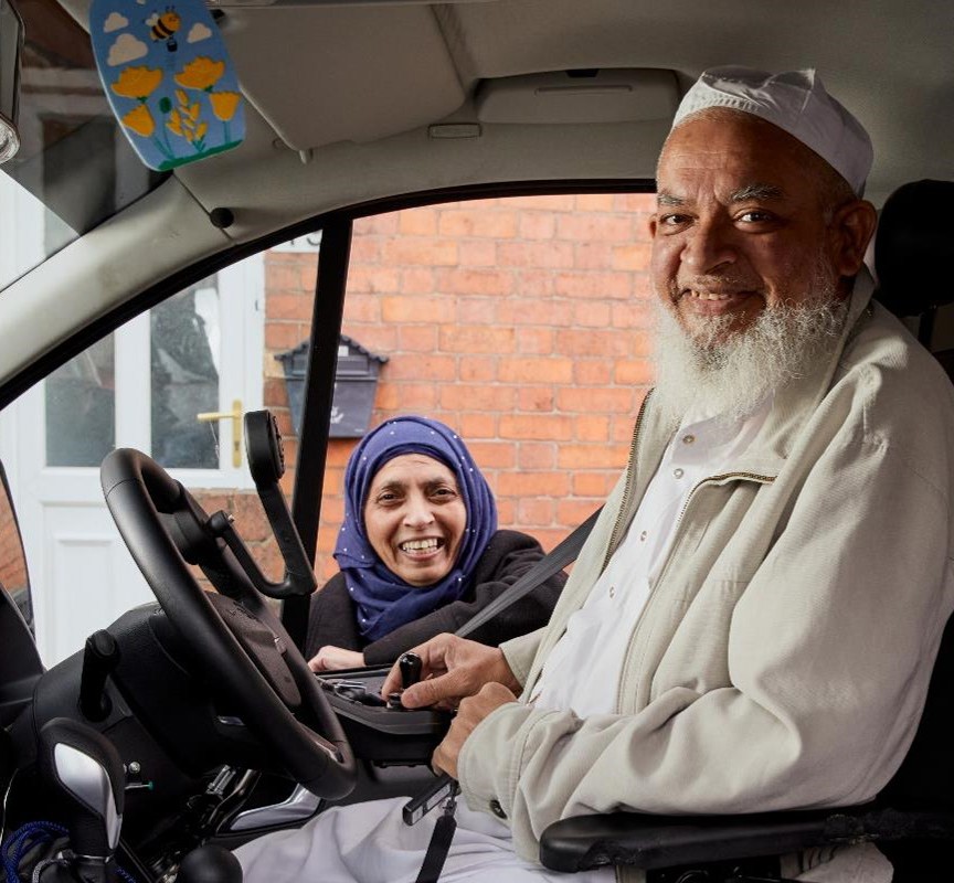 Moosa is sitting in his vehicle with hand controls smiling. His wife is behind the car, standing next to Moosa, smiling. 