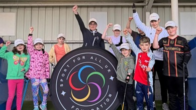 A group of children, young people and adults with limb differences. They are raising on hand in the air. In the middle is a sign that reads: Sport for all UK foundation.