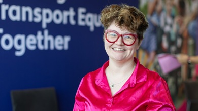 Hannah is smiling to camera, she has short curly brown hair, large red rimmed glasses and a bright pink silk top. 