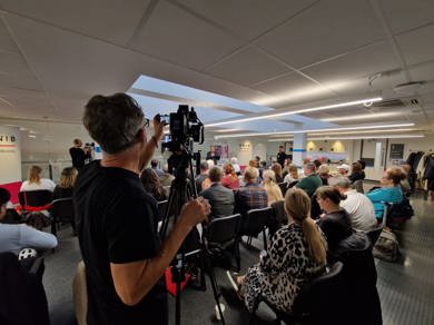 A room of people are sitting on chairs facing the front of the room listening to presentations. A videographer is filming the group. 