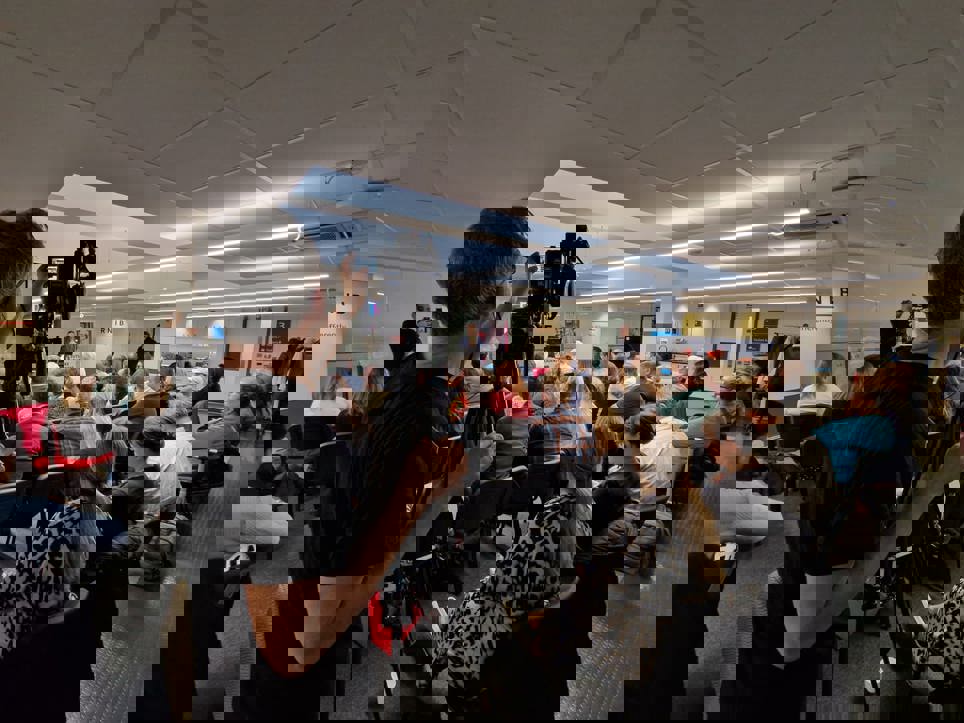 A room of people are sitting on chairs facing the front of the room listening to presentations. A videographer is filming the group. 