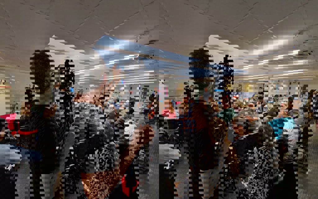 A room of people are sitting on chairs facing the front of the room listening to presentations. A videographer is filming the group. 