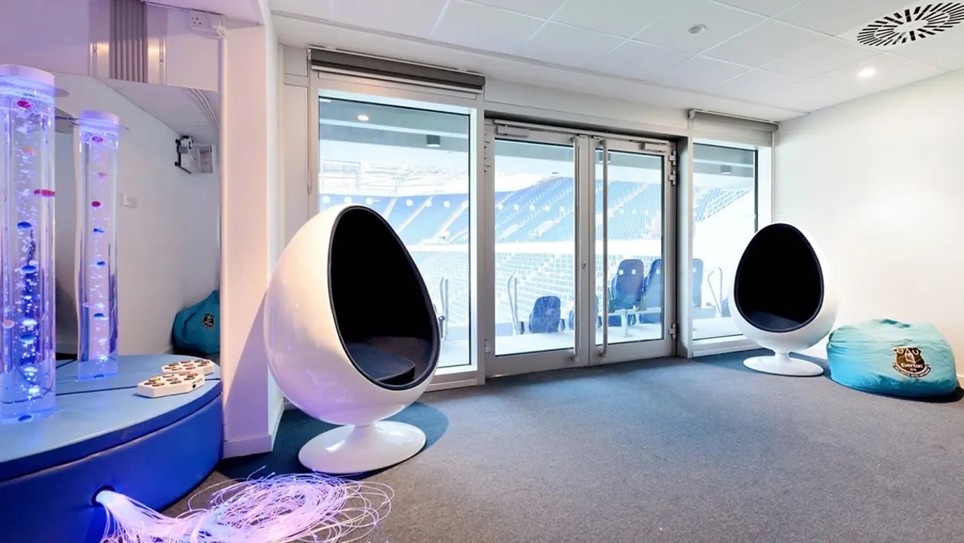Everton Football Club sensory room with white egg pod chairs, bubble tubes, and glass doors overlooking the stadium seating.