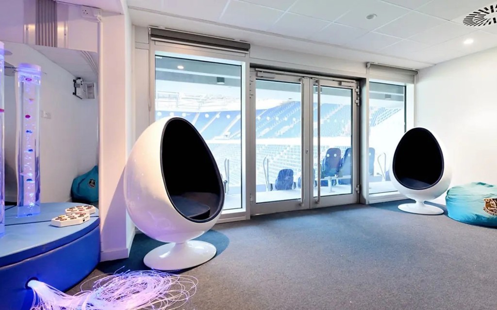 Everton Football Club sensory room with white egg pod chairs, bubble tubes, and glass doors overlooking the stadium seating.