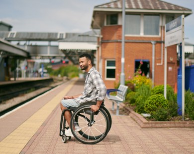 A man with short brown hair is sitting in a wheelchair at a train platform. He is waiting for a train to appear.