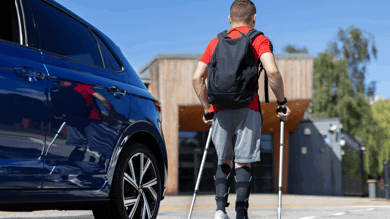 Ben is walking away from his blue vehicles using crutches. He had a red top on with a black backpack and blue shorts. 