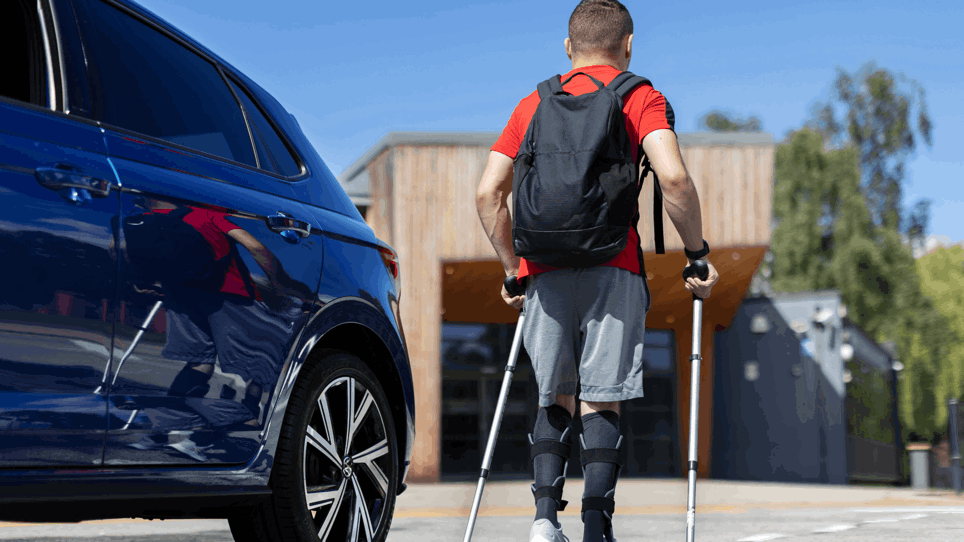 Ben is walking away from his blue vehicles using crutches. He had a red top on with a black backpack and blue shorts. 