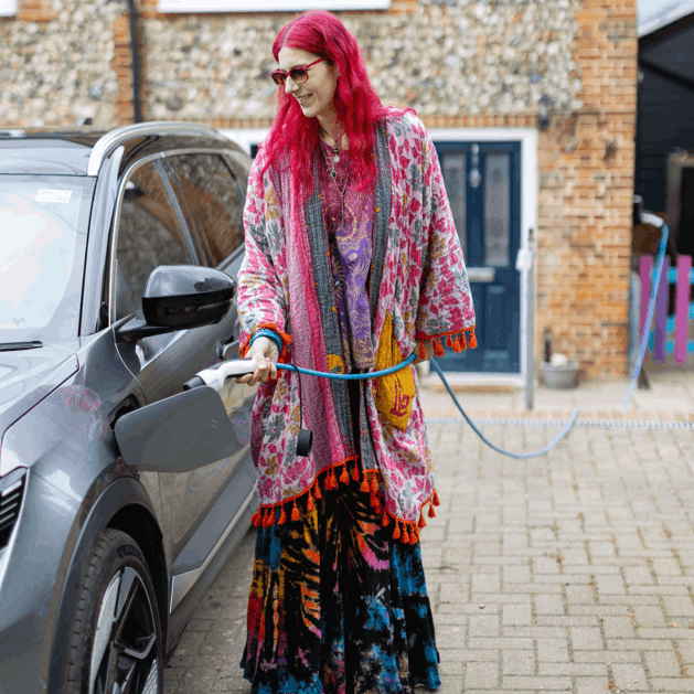 Charlie has pink hair and is wearing multi coloured clothing. They are standing next to their silver vehicle plugging in the electric vehicle charging cable.
