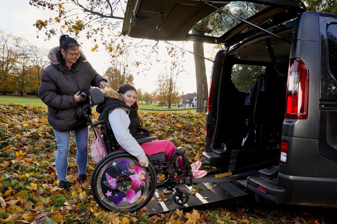 Becky is in her wheelchair on the ramp of her black wheelchair accessible vehicle. She is smiling to camera. She has long brown hair, black coat, grey hoodie, pink trousers and pink trainers. He mum Sarah is operating the ramp, she is has black long hair in a pony tail with a black coat, blue jeans and black trainers, she is smiling at Becky. Behind them is a park with orange and yellow leaves on the trees.