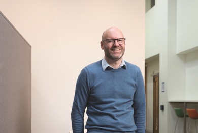 Nigel Fletcher CEO is smiling in a blue jumper with black rimmed glasses. 