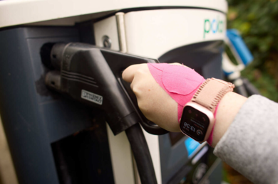 A women is using an EV charging point, she is plugging in a charging cable into electric EV chargepoint. She is wearing a grey coat, pink watch and a pink wrist support.