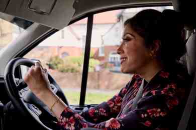 Nazmin is driving her vehicle using a steering wheel hand control. Nazmin has brown long hair in a bun and is wearing a navy shirt with pink flowers. 