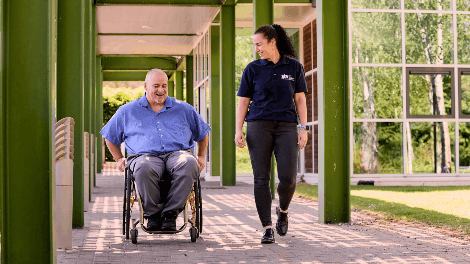 A man is using a manual wheelchair. He is wearing a blue shirt, medium grey trousers and black shoes. A woman walks beside him. She has her wavy hair in a ponytail and is wearing a blue polo shirt with the Spinal Injuries Association logo, dark grey jeans and black shoes.