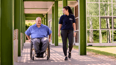 A man is using a manual wheelchair. He is wearing a blue shirt, medium grey trousers and black shoes. A woman walks beside him. She has her wavy hair in a ponytail and is wearing a blue polo shirt with the Spinal Injuries Association logo, dark grey jeans and black shoes.