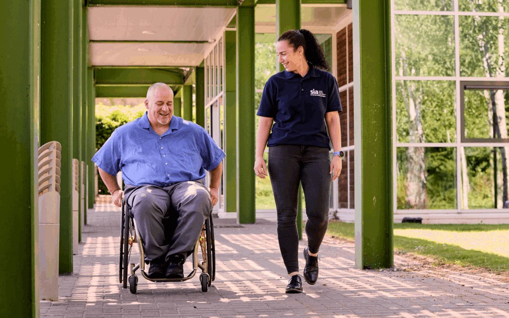 A man is using a manual wheelchair. He is wearing a blue shirt, medium grey trousers and black shoes. A woman walks beside him. She has her wavy hair in a ponytail and is wearing a blue polo shirt with the Spinal Injuries Association logo, dark grey jeans and black shoes.