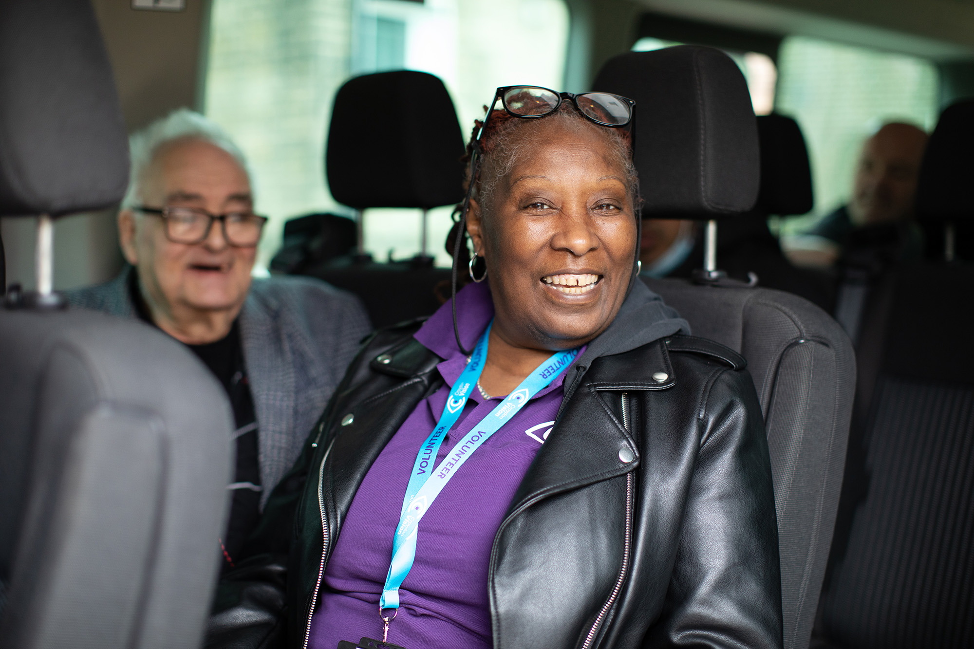 A smiling woman wearing a purple polo shirt sits in a minibus next to a man