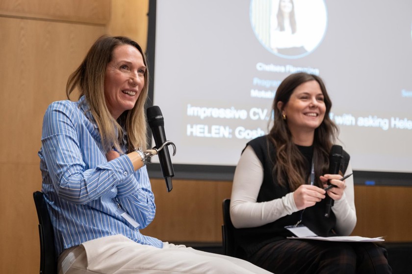 Two women are sitting on chairs at the front of a room, smiling, and presenting with a presentation screen behind them. The first women is wearing a white and blue striped top and the second women is wearing a white and black top. 